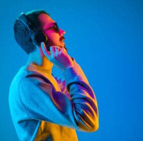 Enjoying his favorite music. Happy young stylish man in sunglasses with headphones listening and smiling while standing against blue neon background